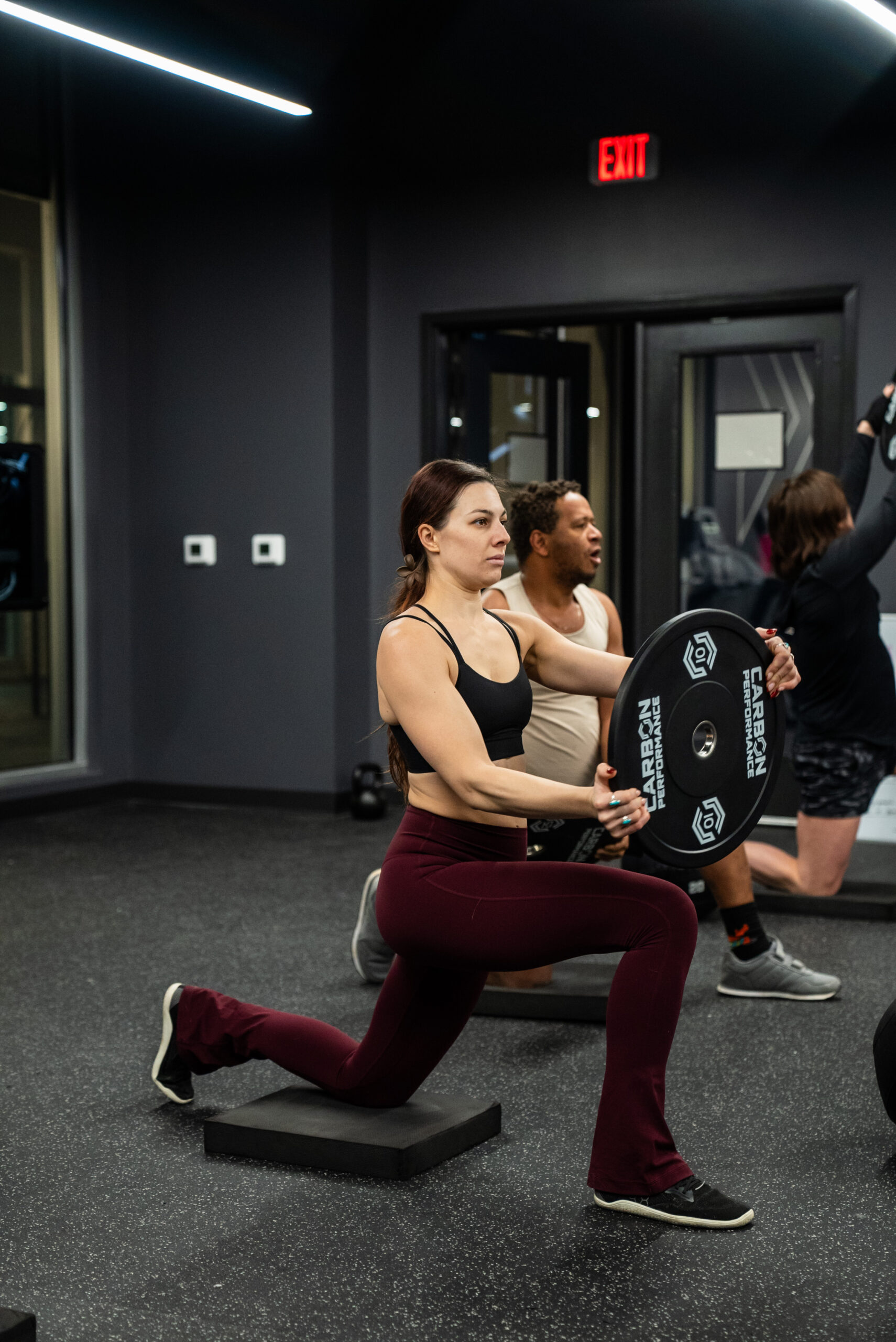 Carbon – February Edge Class-32 A woman lunging with a Carbon Performance weight plate.