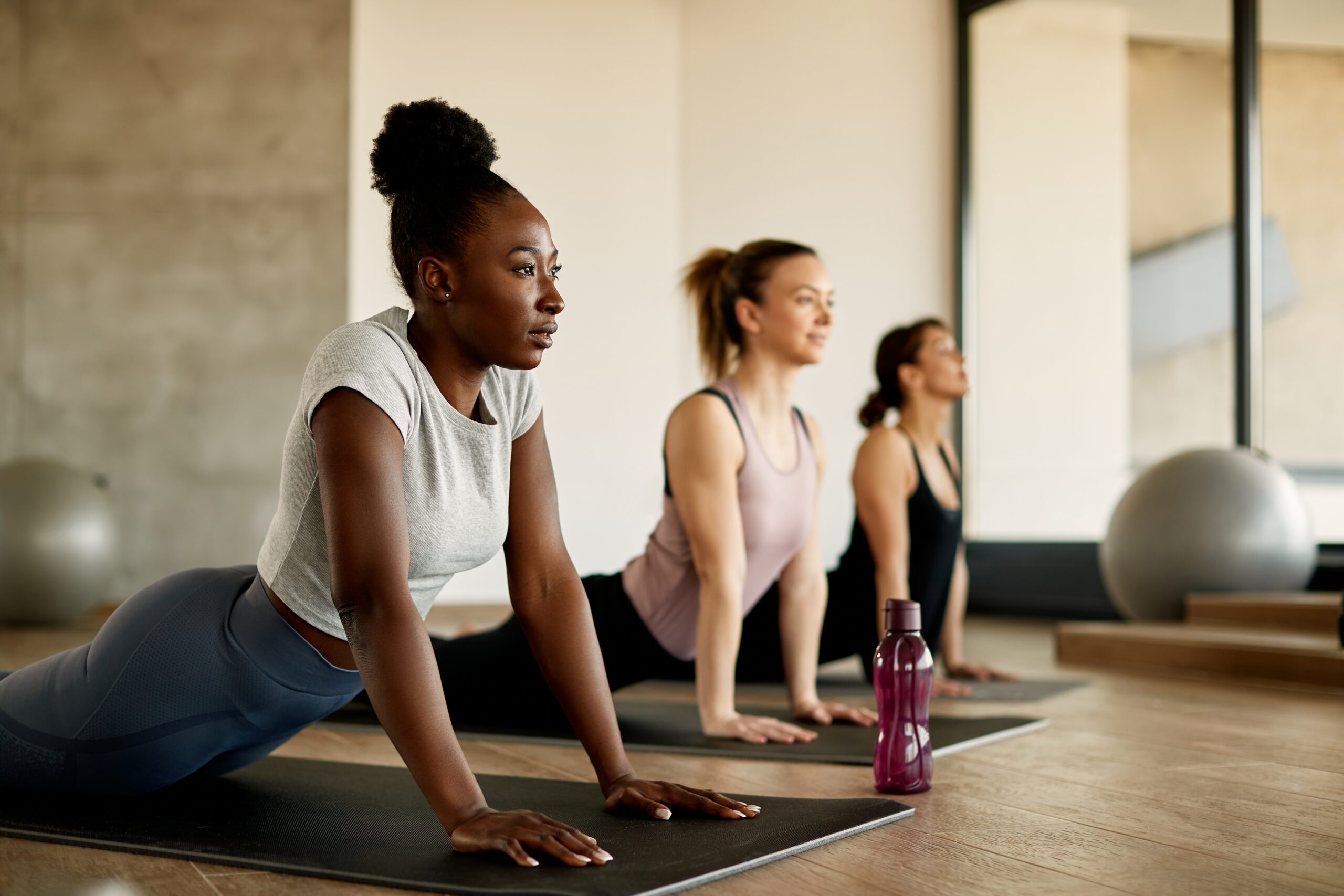 3 women performing yoga pose on mat in sunlit gym.