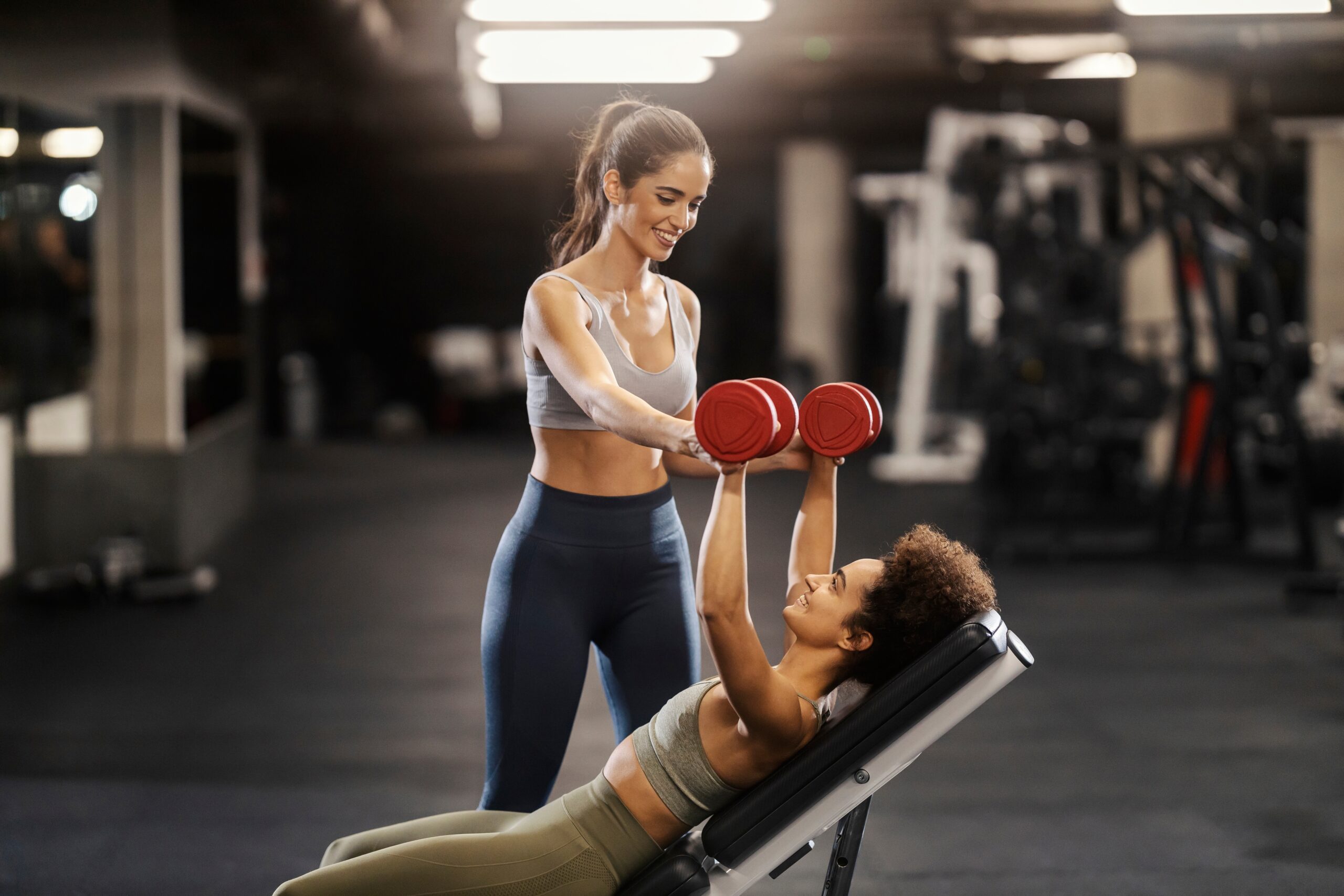 Female personal trainer helping with female lift weights in gym.